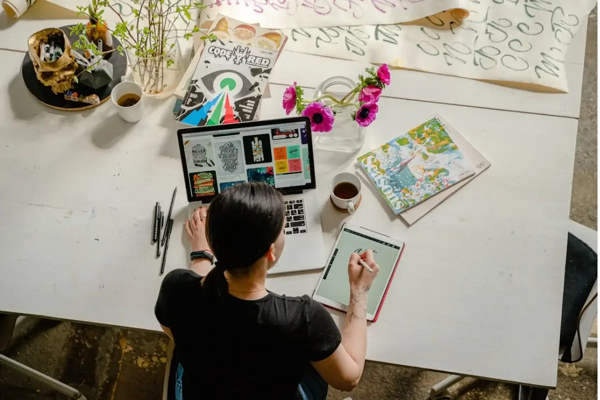 Mujer dibujando en una tableta gráfica, tiene una laptop donde se ven algunas imágenes. Sobre la mesa tiene un café, un florero con flores y algunas hojas