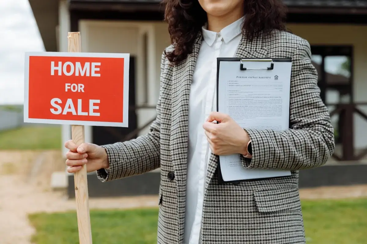 Mujer sosteniendo un aviso de venta de casa con su mano derecha. En la mano izquierda tiene unos documentos