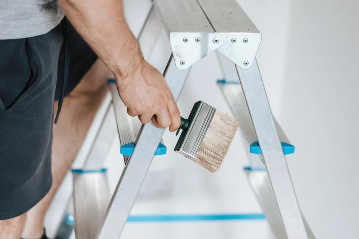 Se ve parte de una persona subiendo una escalera con una brocha para pintura en la mano derecha