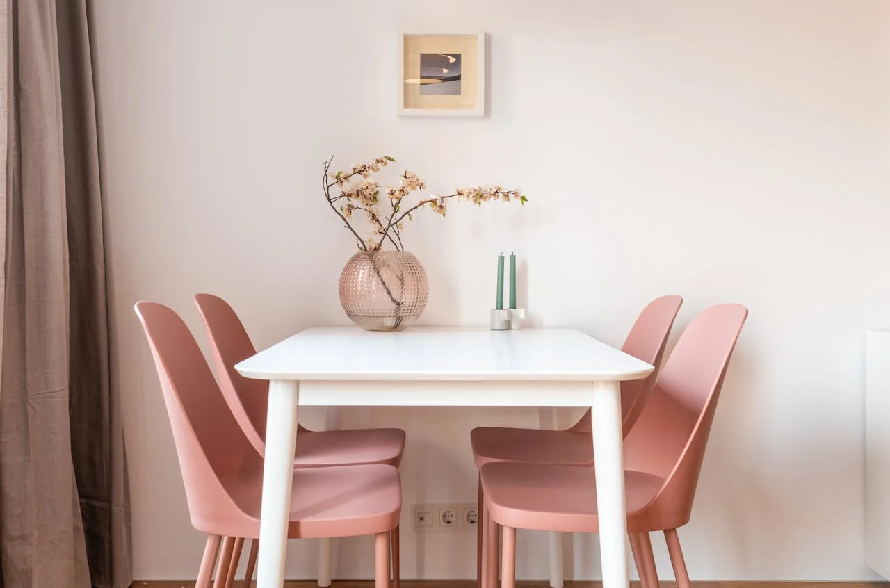 Mesa blanca con 4 sillas color rosa. Sobre la mesa hay un florero con algunas flores y un pequeño cuadro en la pared