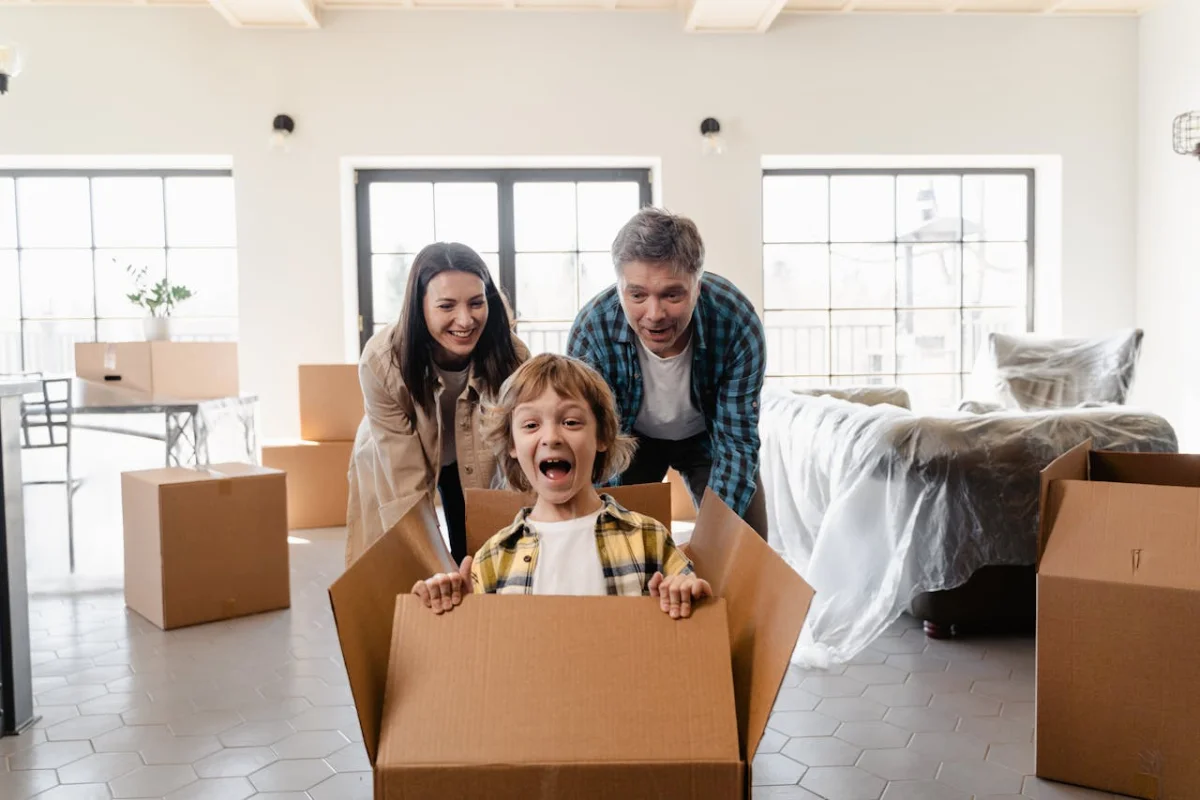 Un hombre y una mujer empujan a un niño dentro de una caja, el niño sonríe. En la habitación en la que se encuentran se ven otras cajas de mudanza, algunos muebles y tres ventanas iluminadas