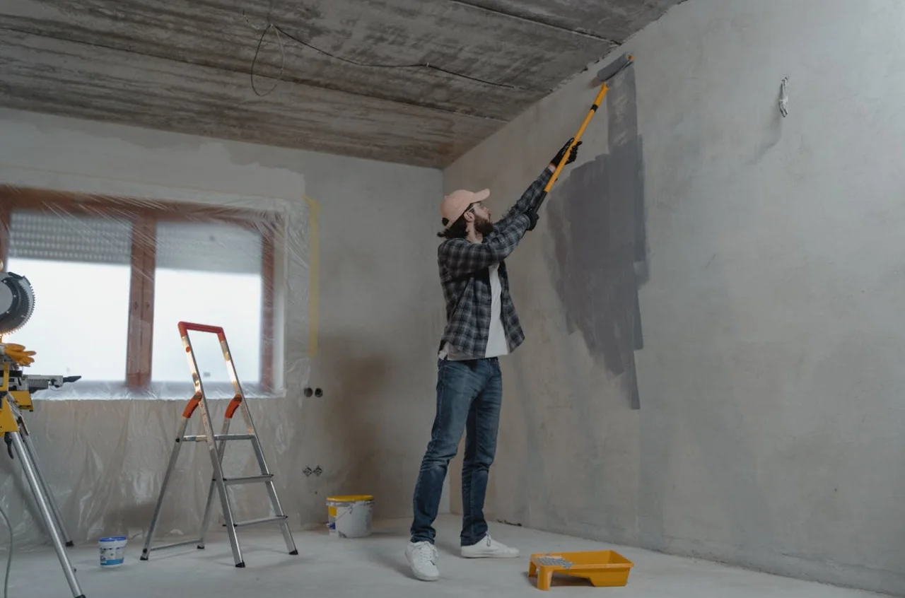 Hombre pintando una pared dentro de una casa, tiene elementos como rodillo, escalera y pintura