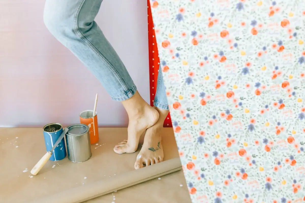 Piernas de una mujer que está de pie en una habitación con tres tarros de pintura, pinceles y un papel tapiz de flores