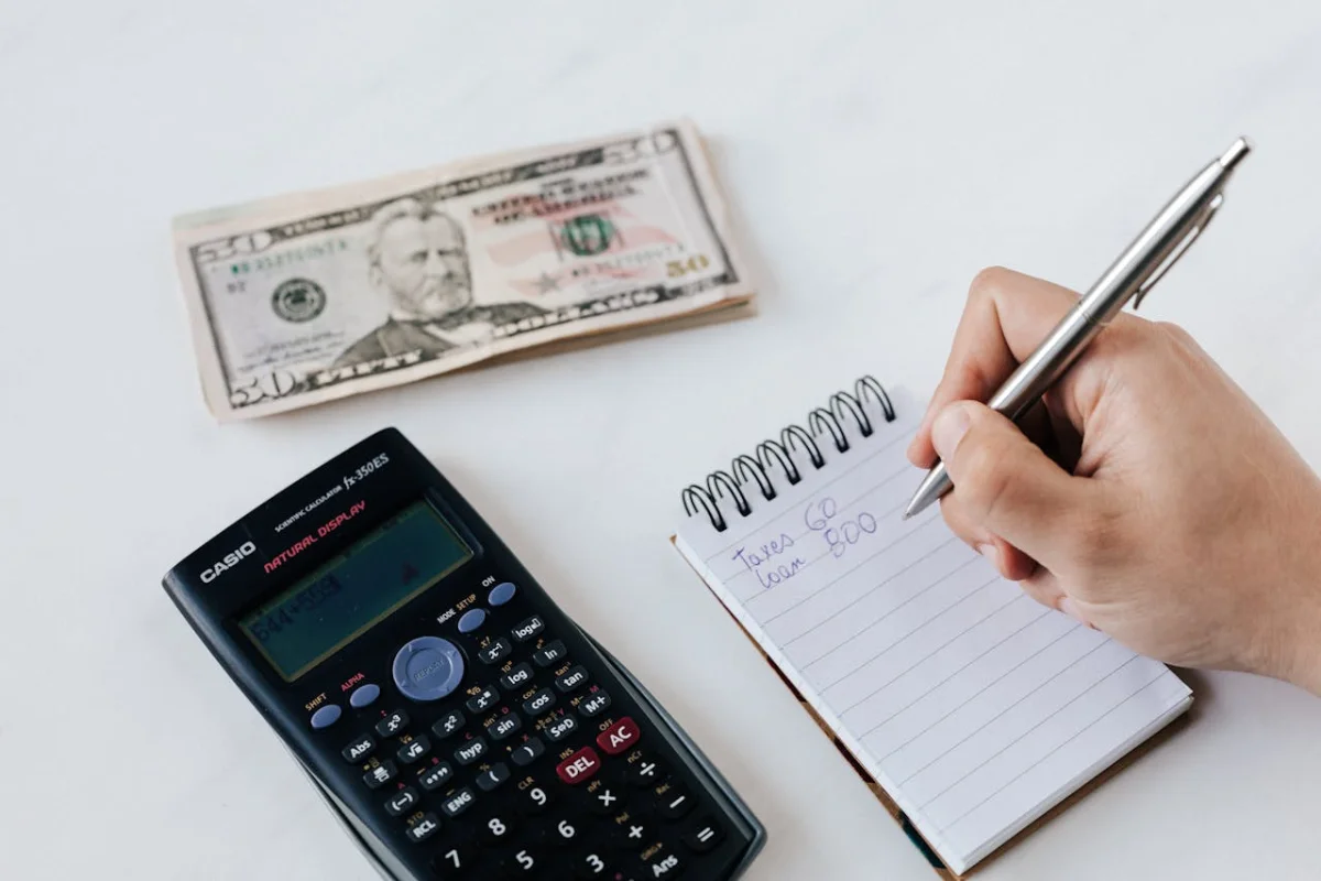 una mano está escribiendo en una libreta, hay unos billetes en frente y una calculadora