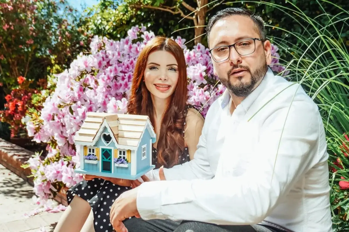 Uno hombre con gafas se encuentra sentado más cerca, a su lado está una mujer son una casa de juguete. Detrás de ellos hay vegetación