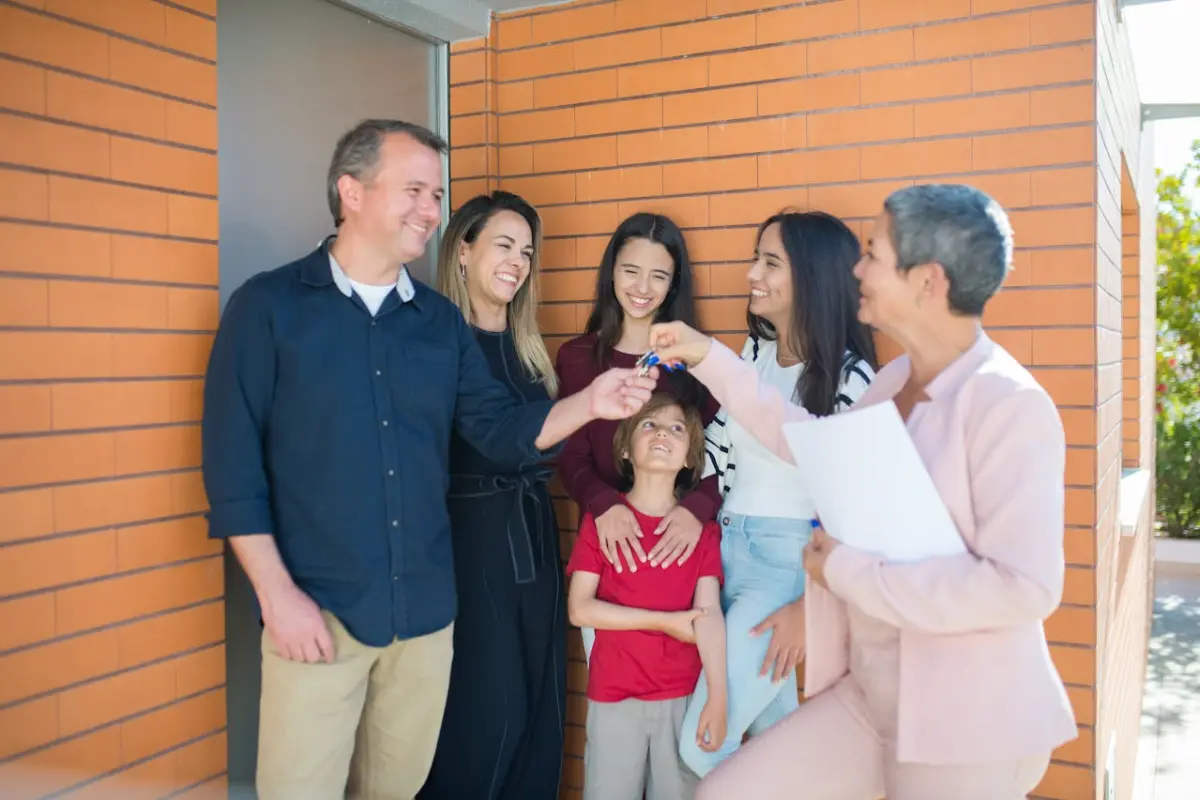 cuatro mujeres, un hombre y un niño. Una de las mujeres tiene unos documentos en su mano y con la otra mano le entrega unas llaves a una de las mujeres. Se encuentran en la puerta de una propiedad inmobiliaria