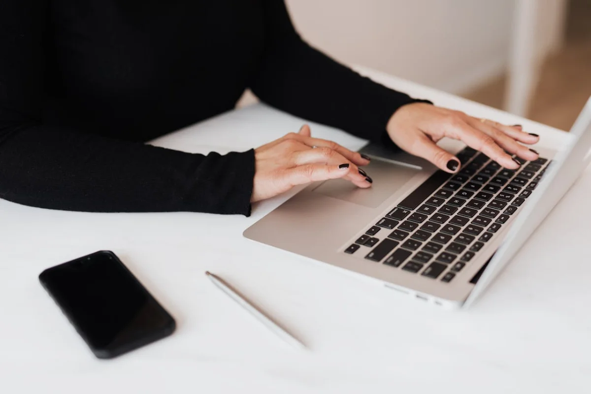 Mano de una mujer escribiendo en una laptop sobre una mesa blanca, a su lado derecho tiene un lapicero y un móvil.