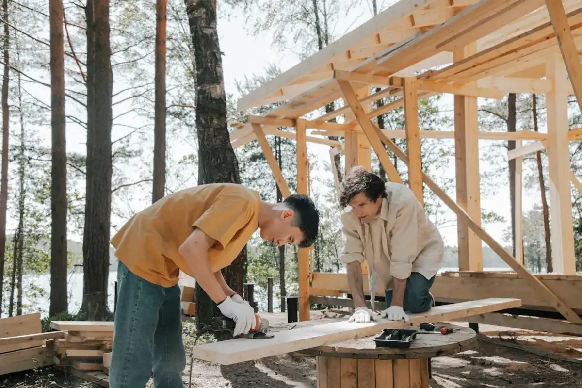 Dos hombres están cortando madera, detrás de ellos se ve la estructura d euna construcción