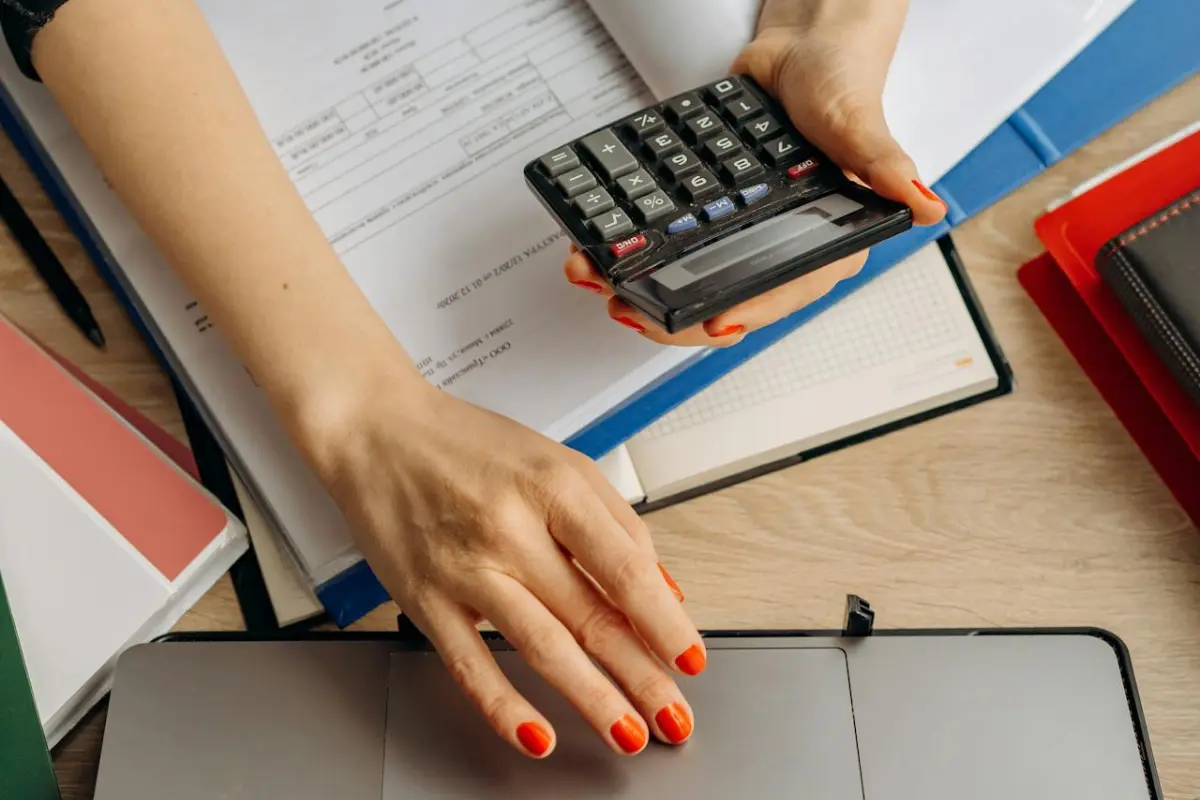 una mujer con una calculadora en su mano y un computador que no se ve del todo. Tiene unos documentos sobre la mesa