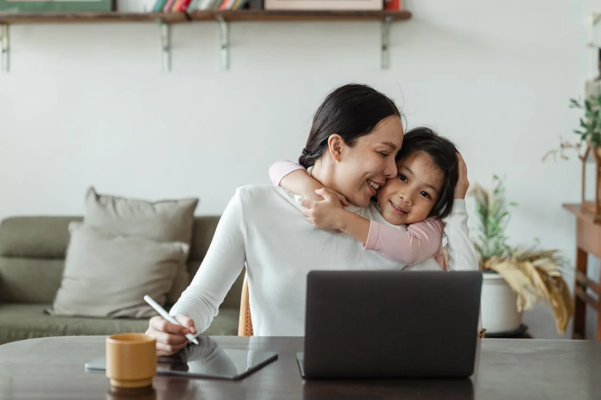 Mujer con una niña que la abraza, se encuentra tomando notas y con una laptop en frente