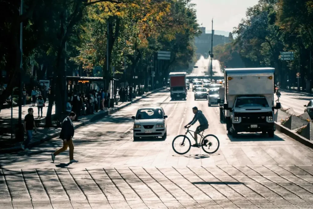 Cale de la CDMX, un hombre va en una bicicleta y otras personas se encuentran caminando o esperando el transporte