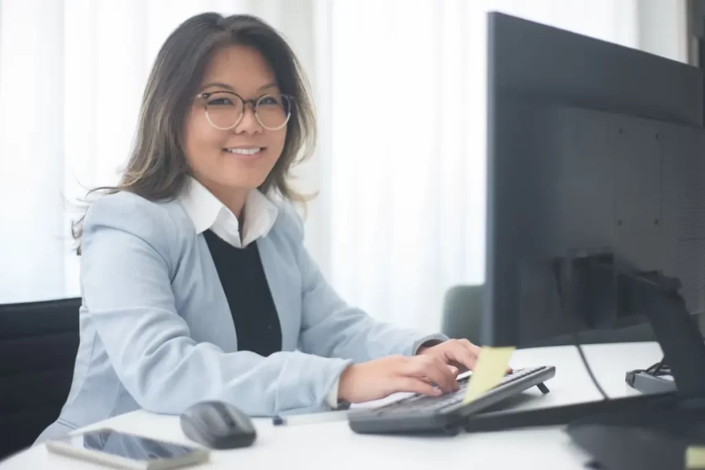 Mujer con gafas sonriendo en una oficina escribiendo en el pc