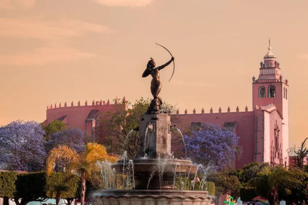 Una fuente con una estatua que tiene un arco. De fondo una estructura arquitectónica grande y algunos árboles