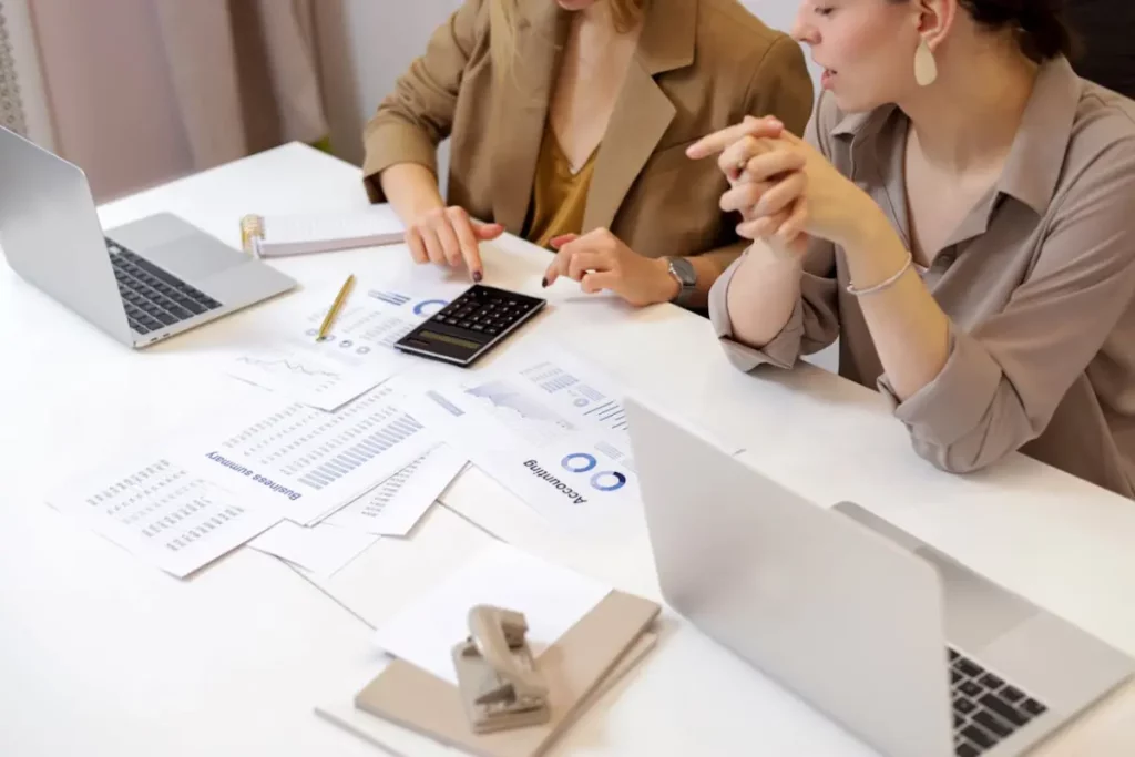 Dos mujeres haciendo cuentas con una calculadora. Tiene gráficas y dos laptops