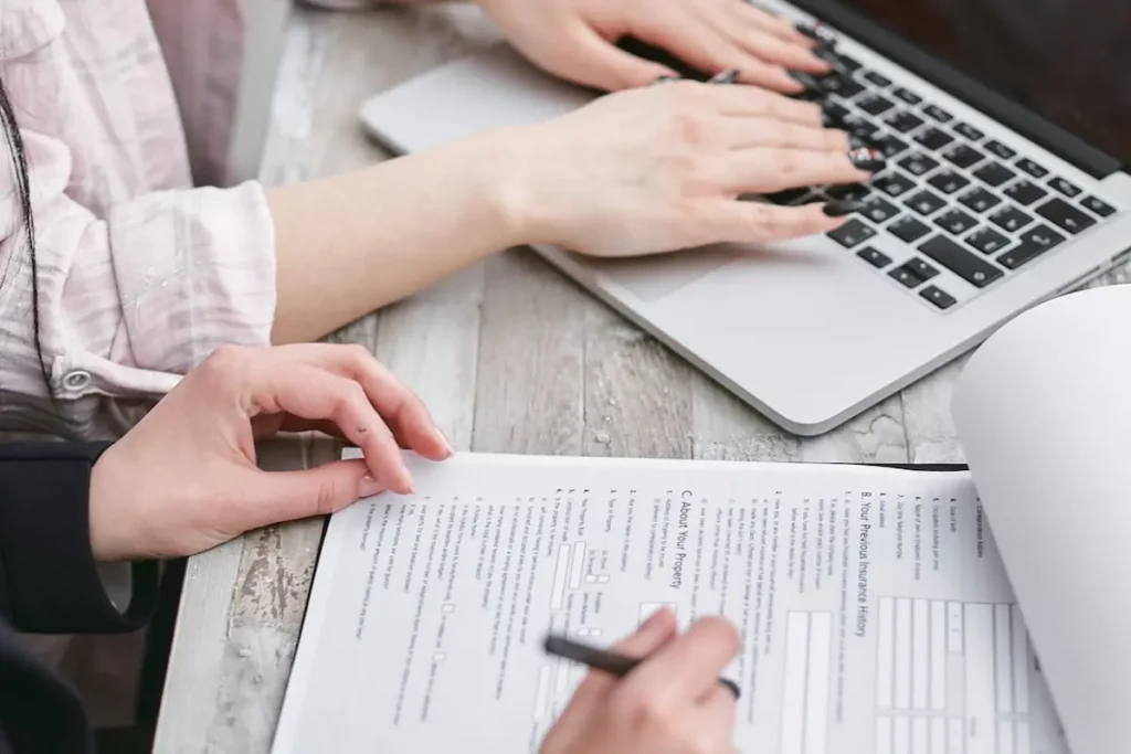 Las manos de dos mujeres, una está escribiendo en una laptop y la otra está escribiendo sobre unos documentos