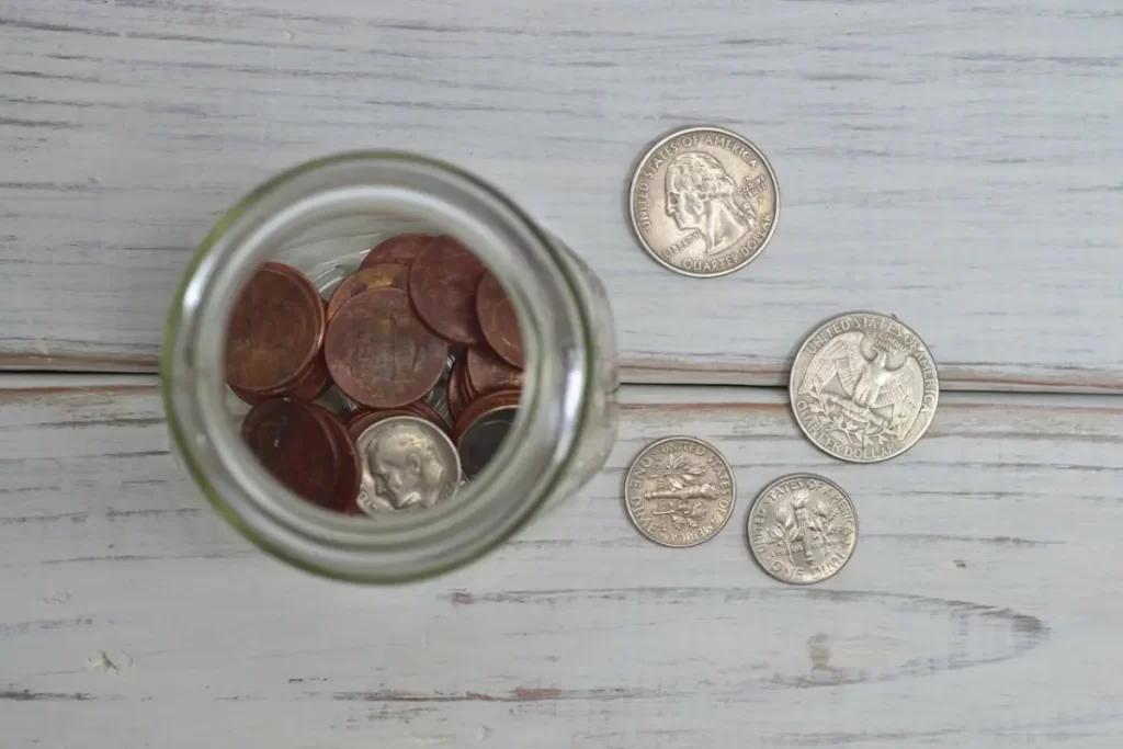 Un frasco de vidrio visto desde arriba con algunas monedas dentro. Se encuentran otras monedas a su lado