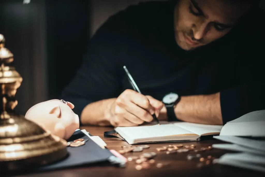 Un hombre escribe en una libreta, tiene monedas y una alcancía en su escritorio