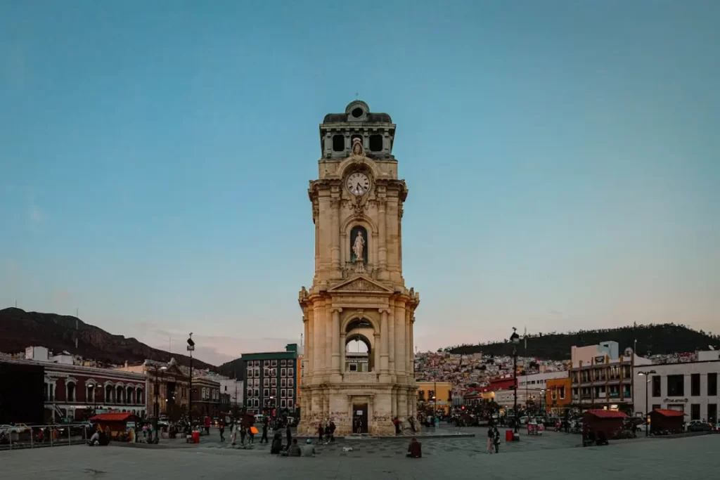 Se ve el cielo, unas montañas y estructuras arquitectónicas con una torre de reloj en el centro de la imagen. Se ven algunas personas