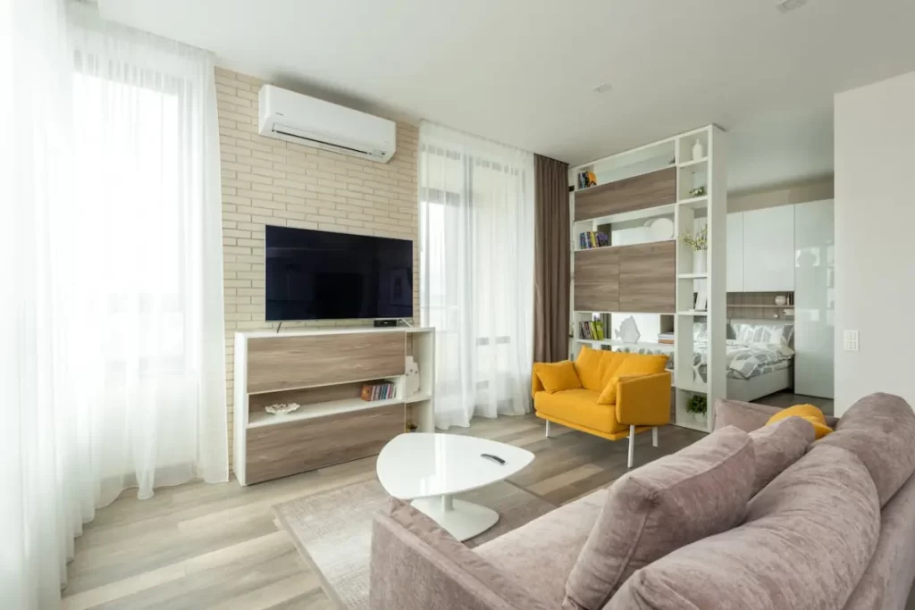 una sala donde está un tv en una repisa con algunos sillones, dos ventanas con cortinas y una estructura que separa este espacio de la habitación principal